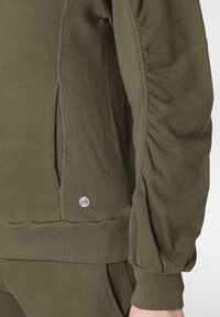 Pullover verde oliva con dettagli sulle maniche arricciate, tessuto morbido, polsini e orlo a costine, dotato di un piccolo logo sul lato inferiore.