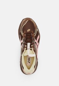 Bruna och beige Asics löparskor med rosa detaljer, mesh överdel, snörning, och vadderad krage sedd ovanifrån på en vit bakgrund.