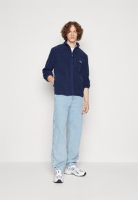 Giacca in fleece navy con colletto alto, zip frontale e due tasche laterali. Il modello indossa jeans azzurri chiari e sneaker bianche. Logo Fila sul petto.