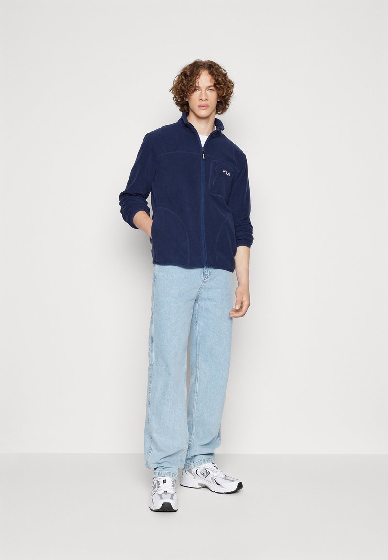 Granatowa polarowa kurtka z wysokim kołnierzem, zamkiem z przodu i dwoma bocznymi kieszeniami. Model ma na sobie jasnoniebieskie dżinsy i białe trampki. Logo Fila na piersi.