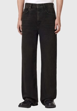 Person iført løse sorte denimjeans og sorte sko, stående mod en lys ensfarvet baggrund.