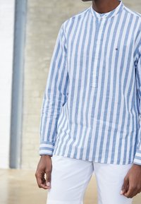 Camicia a maniche lunghe a righe azzurro chiaro e bianco con colletto alla mandarino e pattina con bottoni, realizzata in un tessuto strutturato. Indossata con pantaloni bianchi.