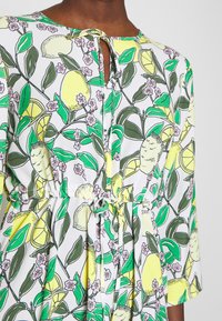 Robe fleurie avec un fond blanc, ornée de citrons jaunes et de feuilles vertes. Comprend un lien à la taille et un décolleté en goutte.