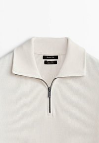 Pull-over blanc côtelé avec un col zippé. Présente un tissu texturé, un léger éclat et une étiquette noire avec le nom de la marque près du col.