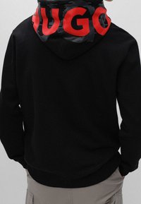 Persona con una sudadera negra con un gran texto rojo "HUGO" en la capucha, combinada con pantalones cargo beis claros, de espaldas.