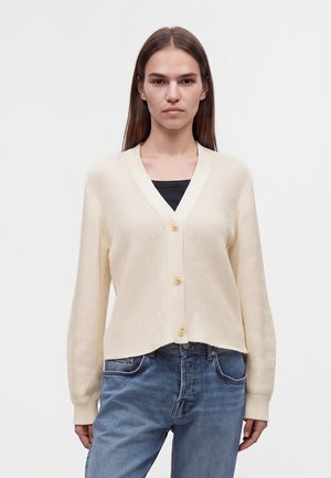 Jeune femme aux longs cheveux bruns, portant un cardigan crème boutonné par-dessus un top noir et un jean bleu, debout devant un fond uni.