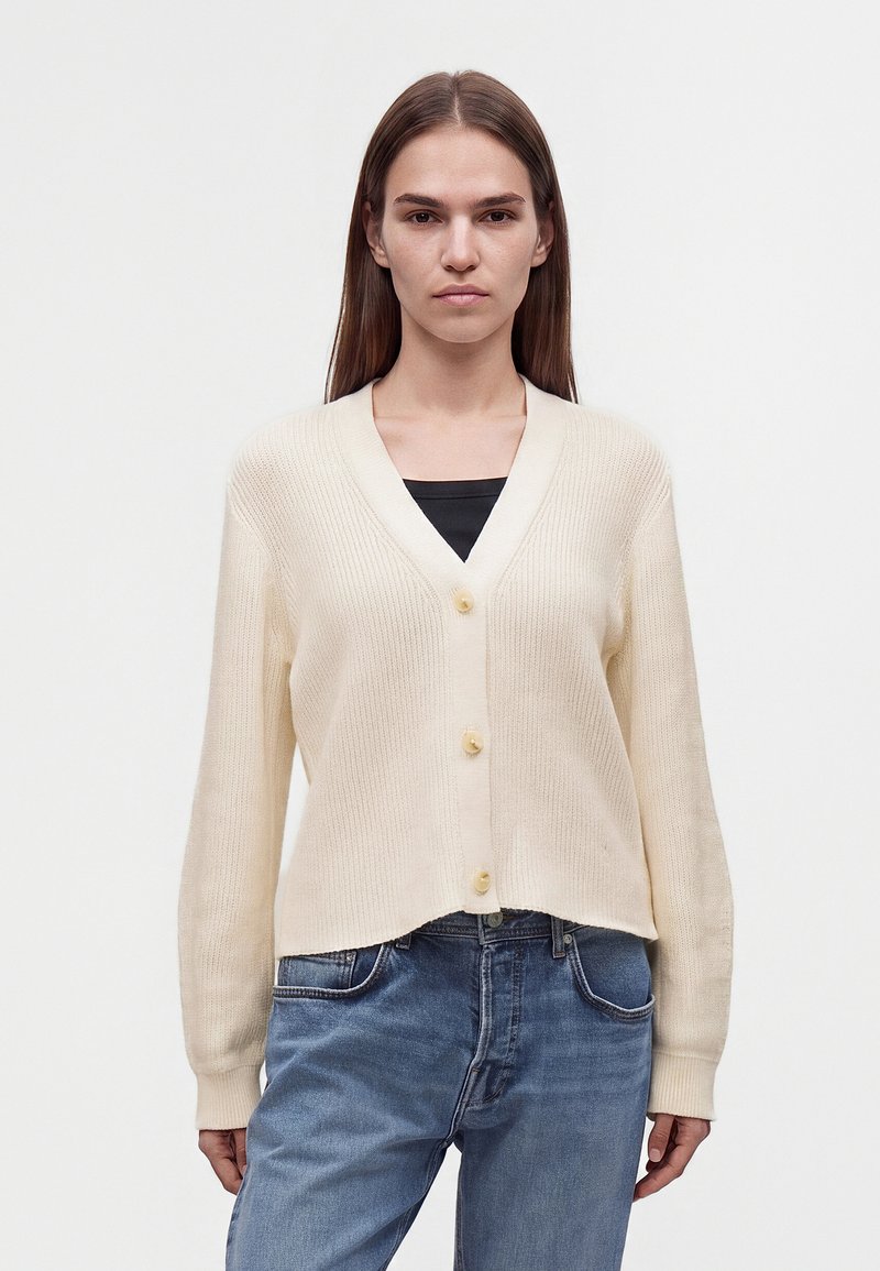 Jeune femme aux longs cheveux bruns, portant un cardigan crème boutonné par-dessus un top noir et un jean bleu, debout devant un fond uni.