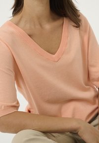 Žena oblečená v svetle broskyňovém svetr s krátkým rukávem a výstřihem do V, sedí s jednou rukou opřenou o koleno.