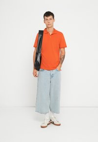 Cămașă polo scurtă, portocalie, blugi largi, bleu deschis, adidași albi cu tălpi maro și o geantă de umăr neagră.