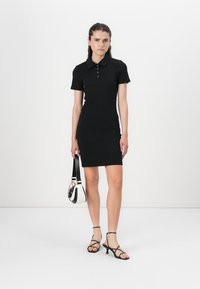 Black ribbed polo dress with short sleeves, fitted shape, three-button placket, paired with black strappy sandals and a small two-tone bag.