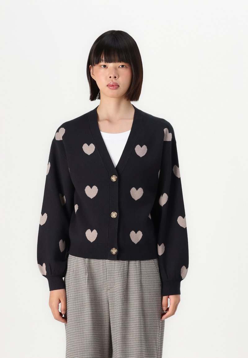 Cardigan blu navy con motivi a forma di cuore marrone chiaro, dotato di scollo a V, chiusura a bottoni e maniche a palloncino. Indossato sopra una maglietta bianca e pantaloni a fantasia.
