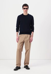 Pull en tricot marine, pantalon beige et chaussures noires. Le modèle se tient droit, portant des lunettes de soleil noires, contre un fond clair uni.