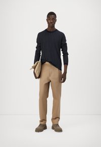 Maglione lavorato a maglia a maniche lunghe blu navy con motivo, pantaloni beige a gamba stretta, scarpe grigie con texture, in mano una borsa a tracolla color nocciola, sfondo semplice.