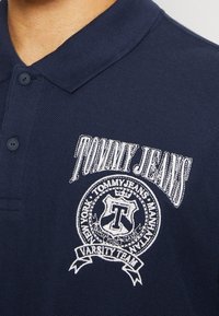 Námořnická modrá polo košile s texturovanou tkaninou, s vyšitým logem "TOMMY JEANS" a kulatým emblémovým designem s akcenty.