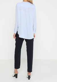 Ljusblå långärmad blus med veck bakåt, kombinerad med marinblå randiga skräddarsydda byxor och vita klackskor med blockklack. Slät tygstruktur.