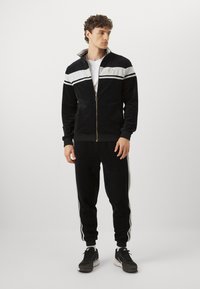 Svart velour treningsdrakt med en jakke med glidelås som har hvite striper og en logo. Tilsvarende striper på tilpassede bukser. Idrettssko påført.
