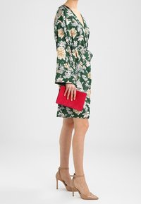 Vihreä kukkakuosilla varustettu pitkähihainen mekko, jossa on V-pääntie, yhdistettynä beigeihin korkokenkiin ja punaiseen clutch-laukkuun. Teksturoitu kuvio, jossa on suuria kukkia.
