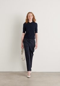 Námořnická polokošile s krátkým rukávem, ve fitted stylu. Tmavé kalhoty, hladká textura. Bílé boty na blokovém podpatku. Modelka nese lehkou kabelku.