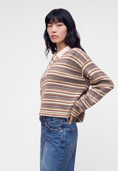 Jeune femme aux cheveux noirs portant une chemise beige rayée à manches longues et un jean bleu, debout avec une main sur la hanche, fond neutre.