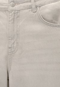 Gros plan sur un jean en denim beige clair montrant la poche avant, la passoire de ceinture et le bouton métallique avec des détails de couture visibles.