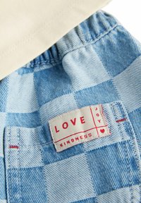 Un short à carreaux bleu clair avec une ceinture élastique et une étiquette affichant "AMOUR" et "BIENVEILLANCE" en texte rouge avec un accent de cœur.