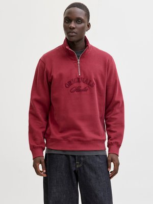 Sweat-shirt rouge avec un col zippé, orné des mots "ORIGINALS" et "Studio" brodés dans un fil assorti. Porté sur un haut gris et un jean sombre.