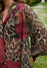 Blouse transparente avec un imprimé floral et animal dans des teintes de rouge, vert et marron, dotée de manches larges et d'un design fluide.