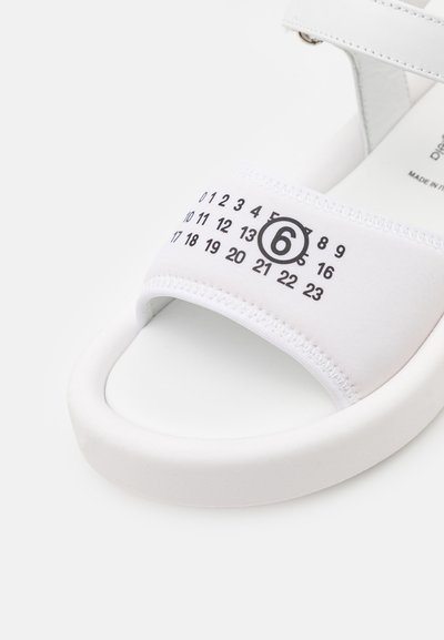 Hvite sandaler med et polstret stoffoverdel og numeriske grafikker trykket i svart. Glatt hvit gummisåle med lett krumning.