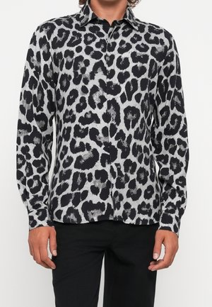 Homme portant une chemise boutonnée à manches longues imprimée léopard noire et grise avec un pantalon noir, debout devant un fond blanc.