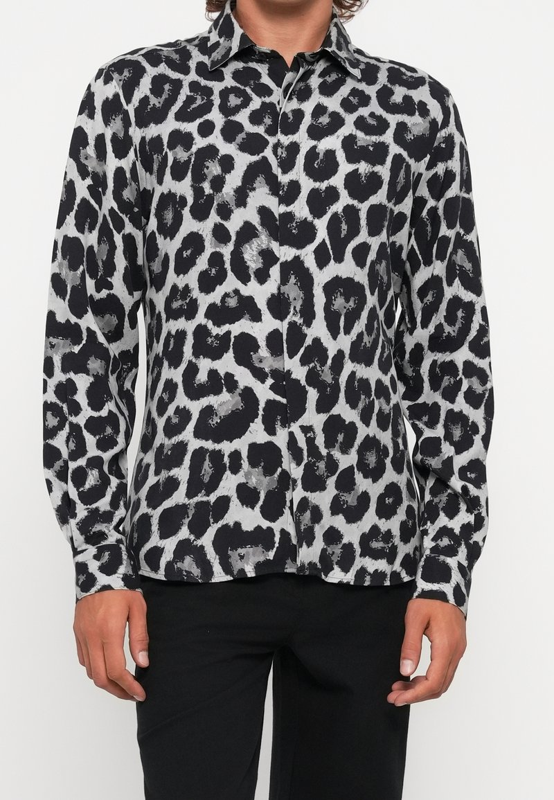 Homme portant une chemise boutonnée à manches longues imprimée léopard noire et grise avec un pantalon noir, debout devant un fond blanc.