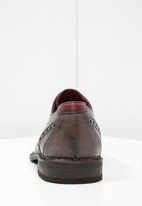 Scarpa di pelle grigia scura con un accento bordeaux sul tallone, caratterizzata da dettagli brogue sui lati e una suola nera strutturata.