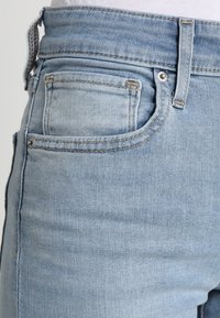 Jeans de mezclilla azul claro con bolsillo frontal visible, presilla para cinturón y sutiles detalles de desgaste en la textura de la tela.