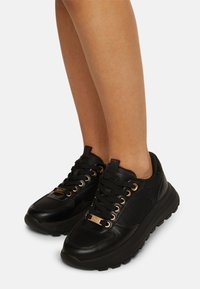 Svarta lädersneakers med gyllene ögla, tjocka sulor och snörning. Har en logodetalj på tungan och en slät yta.