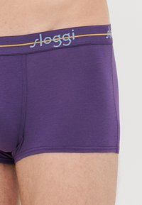 Nahaufnahme einer Person, die lila Sloggi-Boxershorts mit einem blauen und gelben Bund trägt.