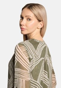 Durchscheinende grüne Bluse mit weißen Streifenmustern und goldenen Akzenten, mit rundem Ausschnitt und weitem Schnitt, aus seitlicher Perspektive gezeigt.