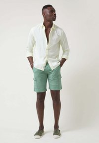 Camisa de lino amarillo claro, desabrochada en el cuello. Pantalones cortos tipo cargo verde menta con bolsillos laterales. Zapatillas grises con suelas blancas.