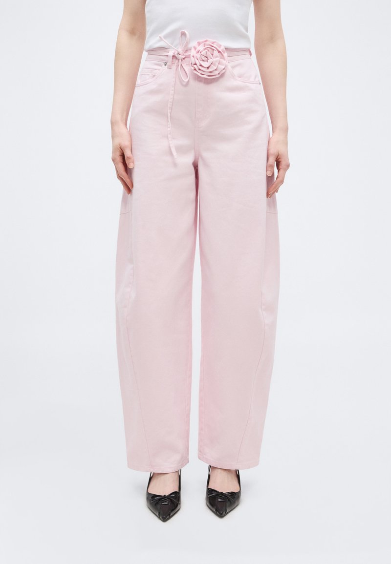 Γυναίκα που φοράει ροζ απαλό παρόμοιο παντελόνι με ψηλή μέση και φαρδιά πόδια, με ζώνη με floral σχέδιο και μαύρες μυτερές γόβες, στέκεται μπροστά από απλό φόντο.