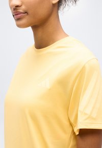 Κοντινό πλάνο ενός ατόμου που φοράει ανοιχτό κίτρινο πουκάμισο Adidas με τρεις αχνές λωρίδες στο μανίκι και λογότυπο στο στήθος.