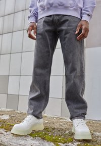 Grå denimjeans med en avslappnad passform, kombinerade med vita skor som har en tjock genomskinlig sula. Omgivningen har betongtexturer och grönska.