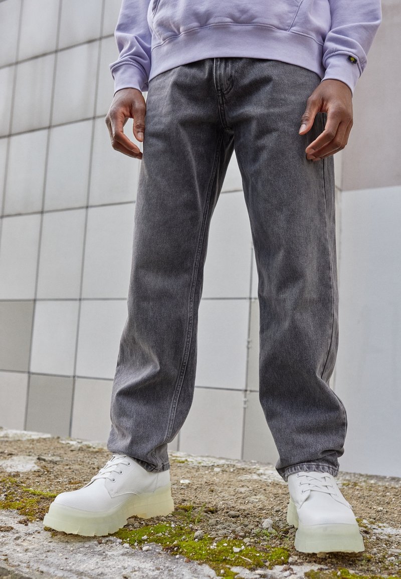 Grå denimjeans med en avslappnad passform, kombinerade med vita skor som har en tjock genomskinlig sula. Omgivningen har betongtexturer och grönska.