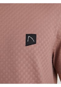 Teksturowana tkanina w kolorze mauve z małą czarną łatą, na której znajduje się biały abstrakcyjny wzór linii na klatce piersiowej krótkiego rękawa.