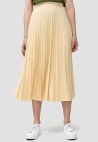Donna che indossa una gonna midi plissettata color giallo chiaro abbinata a scarpe da ginnastica bianche, in piedi di fronte a uno sfondo grigio chiaro uniforme.