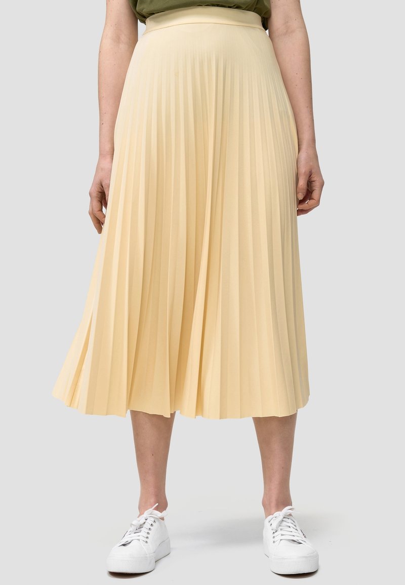 Donna che indossa una gonna midi plissettata color giallo chiaro abbinata a scarpe da ginnastica bianche, in piedi di fronte a uno sfondo grigio chiaro uniforme.