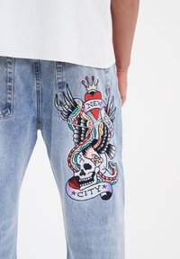 Jeans de mezclilla azul con un colorido diseño bordado de una calavera, una serpiente, alas y el texto "NUEVA YORK" en la pierna izquierda.