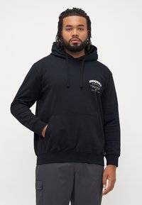 Man with beard and dreadlocks wearing black hoodie with white text on chest and dark gray pants standing with one hand in pocket.