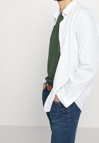 Bílá košile na knoflíky přes zelené tričko, doplněná modrými džíny a hnědým opaskem. Košile má volný střih a rozhalený límec.