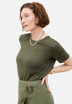 Femme aux cheveux courts et bruns portant un t-shirt vert olive, une jupe assortie avec une boucle circulaire, un collier en chaîne dorée et un bracelet en or.