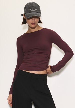 Donna che indossa un cappellino scuro "New York City Brooklyn", una maglietta aderente a maniche lunghe color bordeaux e pantaloni neri a vita alta su uno sfondo bianco.