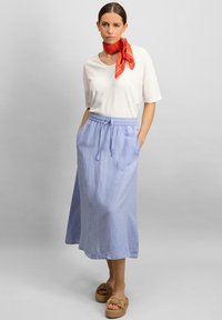Fustă maxi din in de culoare bleu deschis, cu talie ajustabilă cu șnur și buzunare, asortată cu un tricou alb și o eșarfă roșie cu model; sandale din paie cu platformă.