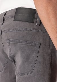 Primer plano de un bolsillo trasero de unos jeans de mezclilla gris y una etiqueta de marca negra en la cintura, llevados por una persona con un brazo visible.